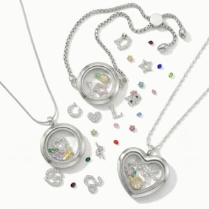 May include: Silver-toned necklaces and bracelet with clear, round lockets. The lockets contain charms like a pineapple, letters, and a heart. Scattered around are various charms, including stars, flowers, and a cat face, all adorned with sparkling crystals.