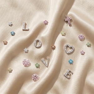 May include: An assortment of jewelry pieces, including the letters L, O, V, and E, along with flower, heart, and cross shapes. The pieces are adorned with sparkling gemstones in various colors, set against a satin background.