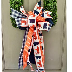 May include: A decorative bow featuring a mix of ribbons in navy blue, white, and orange. The ribbons display patterns of navy blue and white gingham, orange polka dots, and white fabric printed with orange tiger silhouettes and navy blue state outlines.
