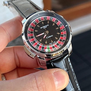 Könnte beinhalten: Eine Luxus-Armbanduhr mit schwarzem Zifferblatt mit einem Roulette-Rad-Design. Die Uhr hat ein silberfarbenes Gehäuse, ein schwarzes Lederarmband und die Marke "JACOB&CO GENEVE" ist sichtbar. Das Wort "CASINO" ist ebenfalls angezeigt.