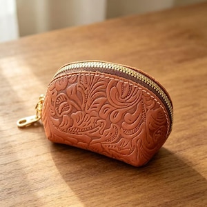 May include: A small, brown leather coin purse with a floral embossed design. The purse has a gold zipper and a gold key ring attachment. The leather is a warm, reddish-brown color.