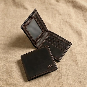 May include: Two brown leather wallets, one open and one closed, displayed on a neutral-toned surface. The open wallet reveals card slots and a clear ID window. The closed wallet features the initials "JM" embossed in the lower right corner. Both wallets have contrasting stitching.