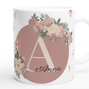 Op de afbeelding: Witte keramische mok met een bloemenontwerp en de letter "A" in een roze cirkel. De naam "Anna" staat onder de letter. De mok is versierd met kleine bloemaccenten.