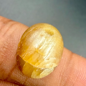 May include: A golden-yellow, oval-shaped gemstone with internal crystalline structures. The polished surface reflects light, showcasing a natural, textured appearance. The gemstone is held in a hand, suggesting its size and portability.