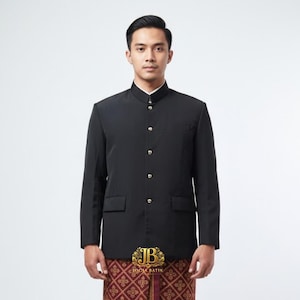 Op de afbeelding: Zwarte jas met een opstaande kraag en gouden knopen. De jas heeft twee voorzakken en wordt gedragen met een patroon sarong. Het logo "JB JOGJA BATIK" is zichtbaar op de sarong.