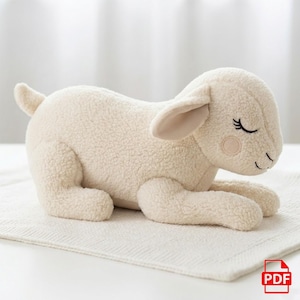 Könnte beinhalten: Ein cremefarbenes Plüschlamm-Spielzeug mit einer weichen, strukturierten Außenseite. Das Lamm liegt mit geschlossenen Augen da, mit schwarzen Wimpern und einer rosa Wange. Das Spielzeug hat beige Innenohren und liegt auf einer weißen, strukturierten Oberfläche.