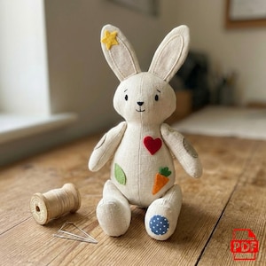 May include: A handmade stuffed bunny rabbit with a cream-colored body, adorned with a red heart, green leaf, and orange carrot applique. The bunny has a yellow star on one ear and blue polka dots on its foot. A wooden spool of thread and pins are in the foreground.
