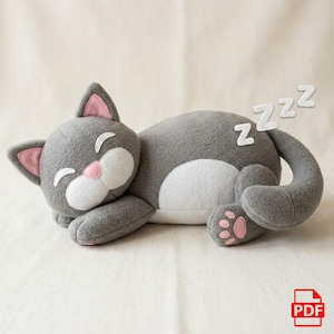 Puede incluir: Un juguete de peluche gris con forma de gato durmiendo, con detalles blancos en la cara y la barriga. El gato tiene el interior de las orejas y las almohadillas de las patas rosas. Letras blancas "Zzz" flotan sobre el gato, indicando sueño.