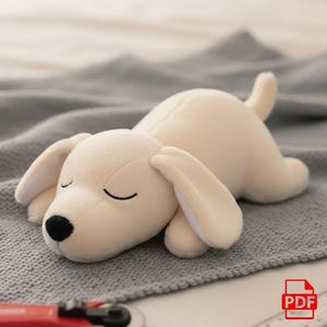 May include: A plush, cream-colored dog toy lying on a gray blanket. The dog has closed eyes, a black nose, and floppy ears. The toy is soft and cuddly, perfect for children or as a decorative item.
