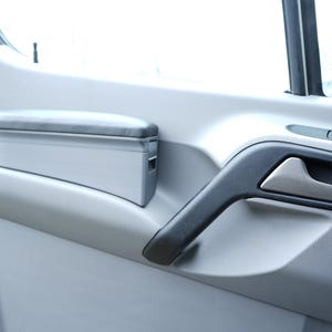 Könnte beinhalten: Innenansicht einer Fahrzeugtürverkleidung. Die Verkleidung ist hellgrau mit einem schwarzen Türgriff und einem silberfarbenen Griff. Eine gepolsterte Armlehne ist an der Türverkleidung montiert.