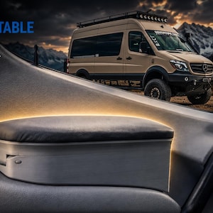 Op de afbeelding: Een donkergrijze gewatteerde armsteun geïnstalleerd op het binnenste deurpaneel van een voertuig, met een zachte kussentop en een voorgevormde basis. De tekst "FIX THE MOST UNCOMFORTABLE PART OF YOUR SPRINTER" is zichtbaar in de linkerbovenhoek.