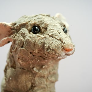 May include: A sculpted figure of a mouse with a rough, textured gray surface. The mouse has prominent, smooth, light pink ears and a small pink nose. Its eyes are round, glossy, and black, providing a sharp contrast to the matte, uneven gray body.