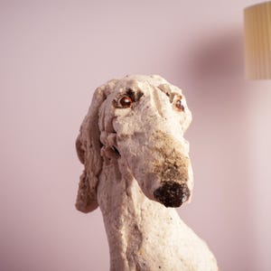 May include: A textured, off-white sculpture of a dog head with long, drooping ears and a prominent snout. The figure features dark, glossy eyes and a dark, mottled nose. The surface has a rough, uneven, hand-crafted appearance with visible clay-like texture.
