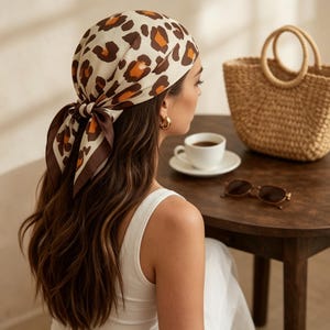 Può includere: Un foulard color crema con stampa leopardata marrone e arancione è legato dietro la testa. Una borsa in vimini, occhiali da sole e una tazza di caffè sono su un tavolo di legno. La scena evoca un'estetica rilassata ed elegante.