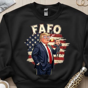 May include: Black sweatshirt featuring a cartoon illustration of a man in a suit smoking a cigar, with the text "FAFO" above. The background includes an American flag design and a smaller cartoon figure.
