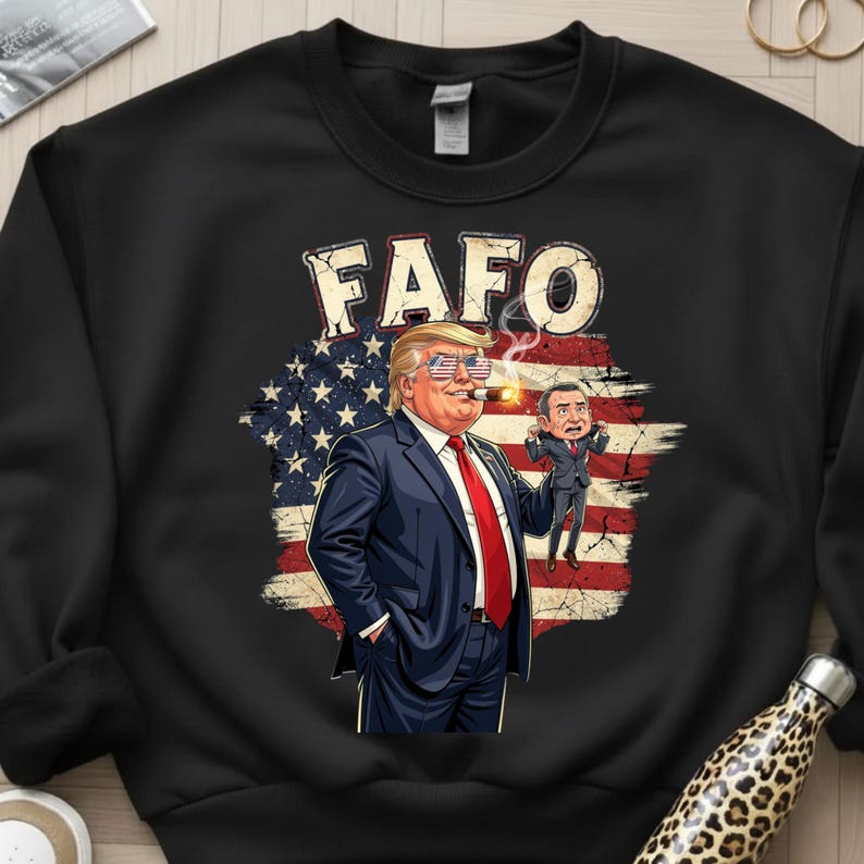 May include: Black sweatshirt featuring a cartoon illustration of a man in a suit smoking a cigar, with the text "FAFO" above. The background includes an American flag design and a smaller cartoon figure.