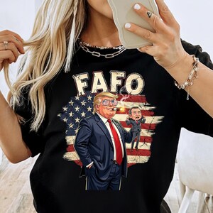 May include: Black t-shirt with the word "FAFO" above a graphic of a man in a suit wearing sunglasses and smoking a cigar, with a smaller figure next to him, set against a distressed American flag background.