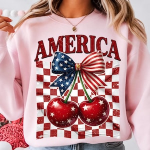 Peut inclure: Un sweat-shirt rose clair à col rond avec une impression graphique de deux cerises rouges à étoiles blanches. Au-dessus des cerises, un grand nœud patriotique à étoiles bleues et rayures rouges sur fond à carreaux rouges et blancs avec le mot AMERICA.