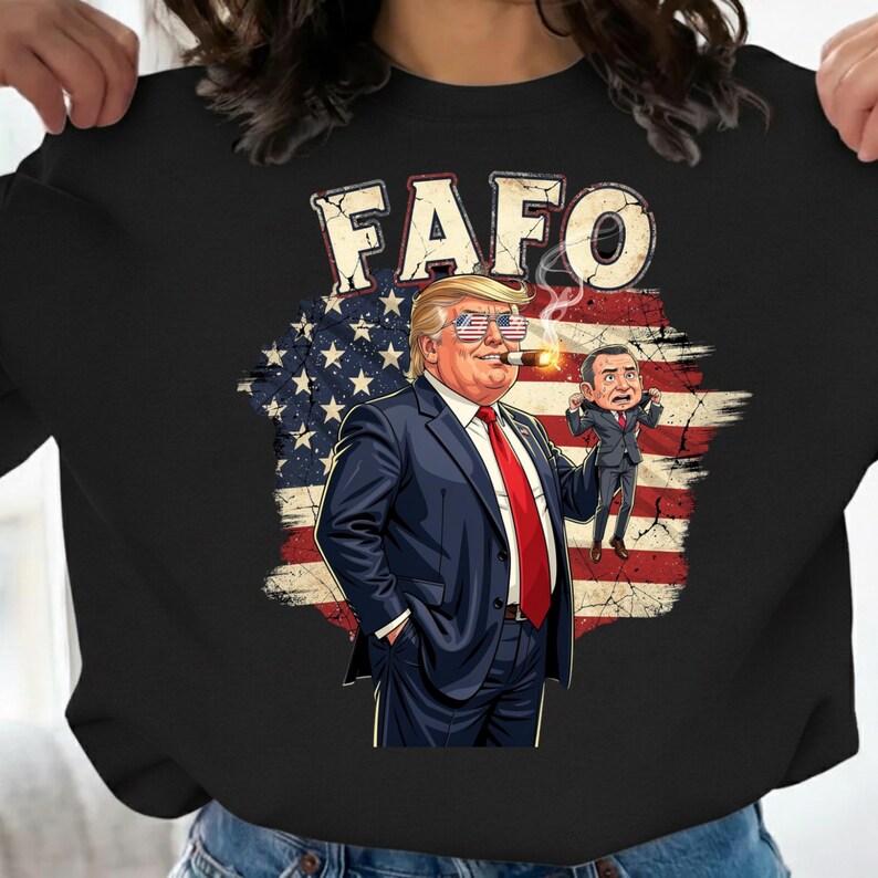 May include: Black sweatshirt featuring a cartoon illustration of a man in a suit, sunglasses, and a cigar, holding a smaller figure. The background includes an American flag design and the text "FAFO" in a distressed font.
