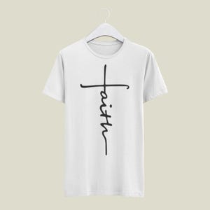 May include: White t-shirt featuring a black cross design with the word "faith" written vertically. The t-shirt is displayed on a white hanger. The design is simple and elegant, suitable for casual wear.