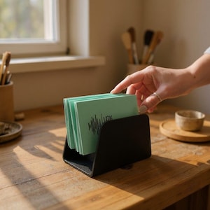 Op de afbeelding: Een zwarte bureau-organizer met een stapel lichtgroene kaarten. Elke kaart heeft een zwarte afbeelding en de letters "1px". De organizer staat op een houten bureau.