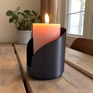 Könnte beinhalten: Eine brennende, pfirsichfarbene Säulenkerze in einem schwarzen Metallhalter. Die Kerze steht auf einem Holztisch, im Hintergrund eine kleine Topfpflanze. Die Kerzenflamme ist hellgelb.