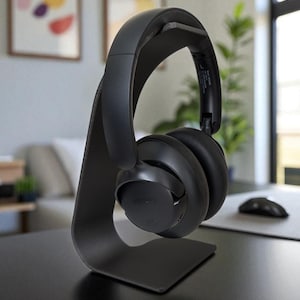 Könnte beinhalten: Schwarzer Over-Ear-Kopfhörer auf einem passenden Ständer. Der Kopfhörer hat ein gepolstertes Kopfband und Ohrmuscheln. Der Ständer ist ein geschwungenes, minimalistisches Design. Der Kopfhörer liegt auf einem schwarzen Schreibtisch, im Hintergrund ist eine Computermaus zu sehen.