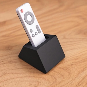 Könnte beinhalten: Eine weiße Fernbedienung mit grauen Tasten und einer roten Ein-/Aus-Taste befindet sich in einem schwarzen, geometrischen Ständer. Der Ständer hat die Form einer Pyramide, und die Fernbedienung ist leicht angewinkelt. Der Hintergrund ist eine Holzoberfläche.