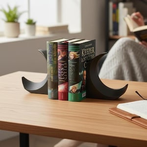 Könnte beinhalten: Drei Bücher, darunter "Harry Potter" und "Keeper of the Lost Cities", werden von einem schwarzen, halbmondförmigen Buchstützen auf einem Holztisch gehalten. Die Bücher haben farbenfrohe Einbände. Im Hintergrund liest eine Person.