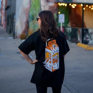 May include: Black t-shirt featuring a milk carton graphic with a skeleton design. The shirt has the words "Meat Intelligence" and "Nutrition Facts" printed on it. A person is wearing the shirt in front of a mural in a street scene.