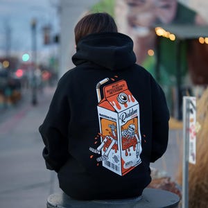 Puede incluir: Sudadera con capucha negra con un gráfico de caja de jugo "Riddim Juice" y una ilustración de esqueleto. El diseño incluye el texto "Beat Intelligence Network" y "Nutrition Facts" en el cartón.
