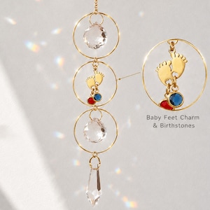 May include: A gold-coloured baby feet charm and birthstone suncatcher. The suncatcher features three gold rings with clear crystal accents and a dangling crystal. The baby feet charms have red and blue birthstone accents. The text "Baby Feet Charm & Birthstones" is visible.
