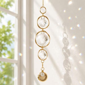 May include: A hanging suncatcher featuring a gold-toned chain with three circular wire frames holding clear faceted crystal spheres. At the bottom, a small gold-toned circular charm displays the letter A and contains a textured beige material.