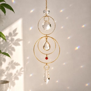 May include: A gold-colored suncatcher with clear crystal accents and a red bead. The suncatcher features a chain, crystal spheres, and circular gold rings. The design is suspended in front of a white wall.