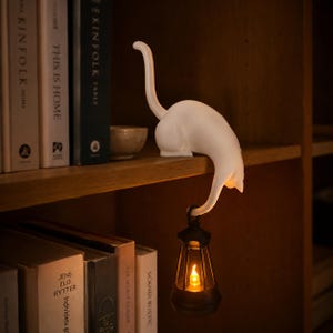 Peut inclure: Une figurine de chat blanc perchée sur le bord d'une étagère avec la queue courbée vers le bas, tenant une petite lanterne noire qui émet une lumière orange chaude et brillante. Le chat est en position accroupie, le dos arqué et la queue levée.