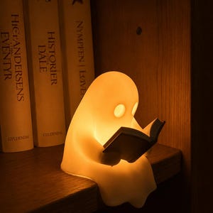 Può includere: Una piccola lampada luminosa a forma di fantasma color bianco sporco, realizzata in materiale liscio, raffigurata seduta su un bordo mentre tiene e legge un libro aperto. La lampada emette una luce arancione calda e soffusa che illumina la forma.