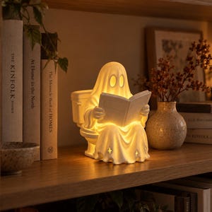 Könnte beinhalten: Eine weiße, geisterförmige Keramiklampe, die auf einer Toilette sitzt und ein Buch liest. Die Lampe strahlt ein warmes, sanftes Licht aus, das die ovalen Augen des Geistes und die sternförmigen Ausschnitte am Saum des Gewandes hervorhebt.