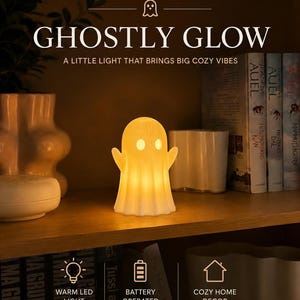 Peut inclure: Une lampe LED blanche en forme de fantôme émettant une lueur chaude. L'image contient le texte GHOSTLY GLOW, A LITTLE LIGHT THAT BRINGS BIG COZY VIBES, WARM LED LIGHT, BATTERY OPERATED et COZY HOME DECOR. La lampe est posée sur une étagère en bois.