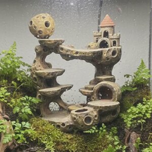 May include: A tan, porous, multi-level resin structure featuring a small castle with a conical roof, a spherical moon-like orb, and several flat platforms, nestled within a terrarium environment surrounded by green ferns and mossy soil.