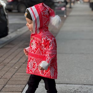 Puede incluir: Un niño con un abrigo de invierno con estampado rojo y blanco y una capucha a juego con un pompón blanco. El abrigo tiene un diseño geométrico detallado. El niño también lleva pantalones negros, un gorro verde y zapatillas grises.
