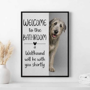 Pode incluir: Uma impressão emoldurada com o texto "WELCOME to the BATHROOM" e "a Wolfhound will be with you shortly." Um cão Wolfhound cinzento espreita por trás do texto. A impressão está numa moldura preta.