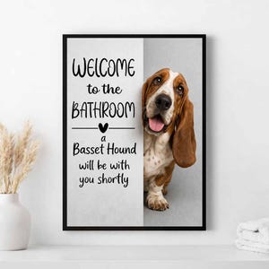 Könnte beinhalten: Gerahmtes Wandbild mit einem Basset Hound Hund mit braun-weißem Fell. Der Text "WELCOME to the BATHROOM" und "a Basset Hound will be with you shortly" ist in schwarzer Schrift geschrieben. Der Rahmen ist schwarz.