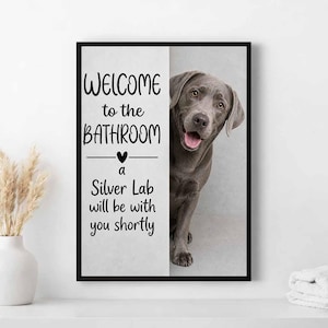 Peut inclure: Tableau encadré avec un Labrador argenté souriant. Le texte dit : « BIENVENUE dans la SALLE DE BAINS », suivi de « un Labrador argenté sera bientôt avec vous ». Le cadre est noir.