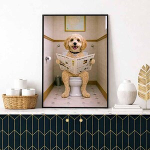 Könnte beinhalten: Ein gerahmter Druck zeigt einen Hund auf einer Toilette, der eine Zeitung liest. Der Hund ist hellbraun und trägt ein schwarzes Halsband. Das Badezimmer hat eine neutrale Farbgebung, mit einem Korb mit Toilettenpapier im Vordergrund.