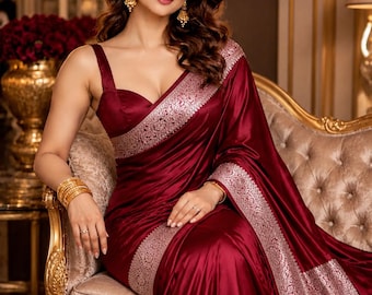 May include: A deep burgundy satin saree featuring a wide, intricate silver metallic border with floral patterns. The garment includes a sleeveless bodice with a sweetheart neckline, paired with gold-toned drop earrings and stacked gold bangles.