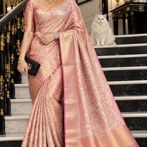 Puede incluir: Un sari de seda rosa y dorado con un intrincado patrón de brocado floral en toda la tela. La prenda incluye un borde ancho de color oro metálico con un ribete rosa fino, mangas cortas y un pallu a juego drapeado sobre el hombro.