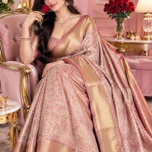 Puede incluir: Un sari de color rosa empolvado con intrincados patrones florales de brocado metálico dorado en toda la tela. La prenda incluye un borde dorado ancho y ornamentado con detalles de rayas diagonales y una blusa a juego de manga corta.