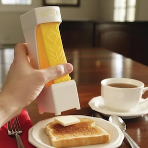 Op de afbeelding: Een witte en gele boterdispenser met het woord "BUTTER" erop gedrukt. Een klontje boter ligt op een sneetje toast op een wit bord. Een kopje thee en bestek staan ook op tafel.