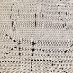 Puede incluir: Un tejido de ganchillo beige con un patrón repetitivo de diamantes, copas de vino y botellas. El diseño incluye las letras K y K formadas por puntos calados, con una gran forma de cuenco en el borde inferior de la tela.