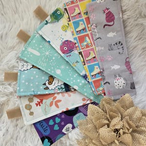 May include: A collection of handmade fabric bookmarks with various patterns. Designs include roller skates, cats, fish, sheep, and a purple bookmark with the word "cute". Each bookmark has a beige fabric tab attached.